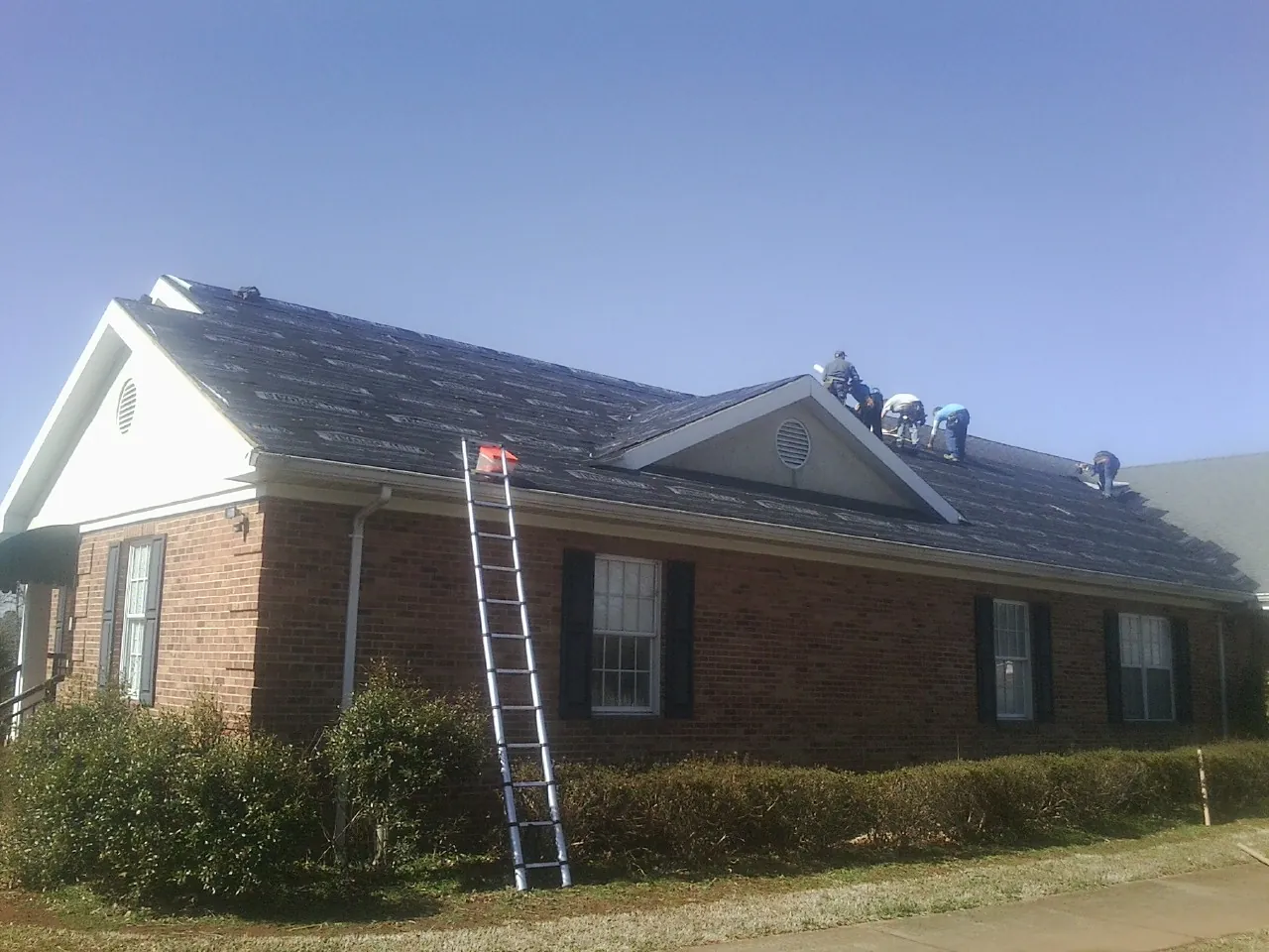 Crew tearing shingles off a brick ranch roof with a ladder set — Metal Roof Repair work by our crew in Ganado