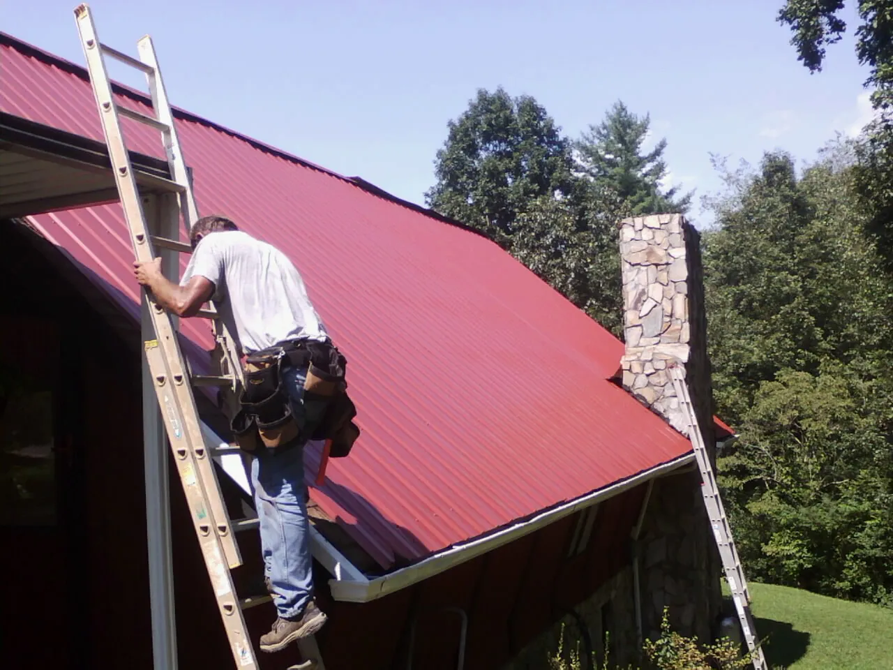Roofer on a ladder beside a red metal roof and stone chimney — Soffit & Fascia Repair by Roofing Brothers in Ganado